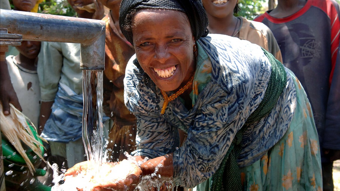 Eine Frau steht an einer Wasserstelle und lacht in die Kamera