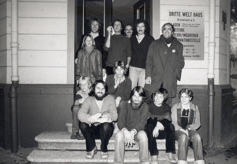 Ein schwarz-weiß-Foto aus den frühen 1980er Jahren zeigt zwölf junge Menschen posierend für die Kamera, davon fünf weibliche und fünf männliche. Sie sitzen und stehen auf den Eingangsstufen zum heutigen Café Welthaus. Ein Schild mit den Öffnungszeiten hängt an der Außenwand, auf dem "Dritte Welt Haus Bielefeld e.V." steht sowie die Angebote Laden, Teestube, Archiv, Mediothek und Informationsstelle.