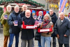 Eine Gruppe Menschen steht vor einem Stand auf dem Weihnachtsmarkt und hält Bauchläden, in denen sich Lotterielose befinden, in ihren Händen.
