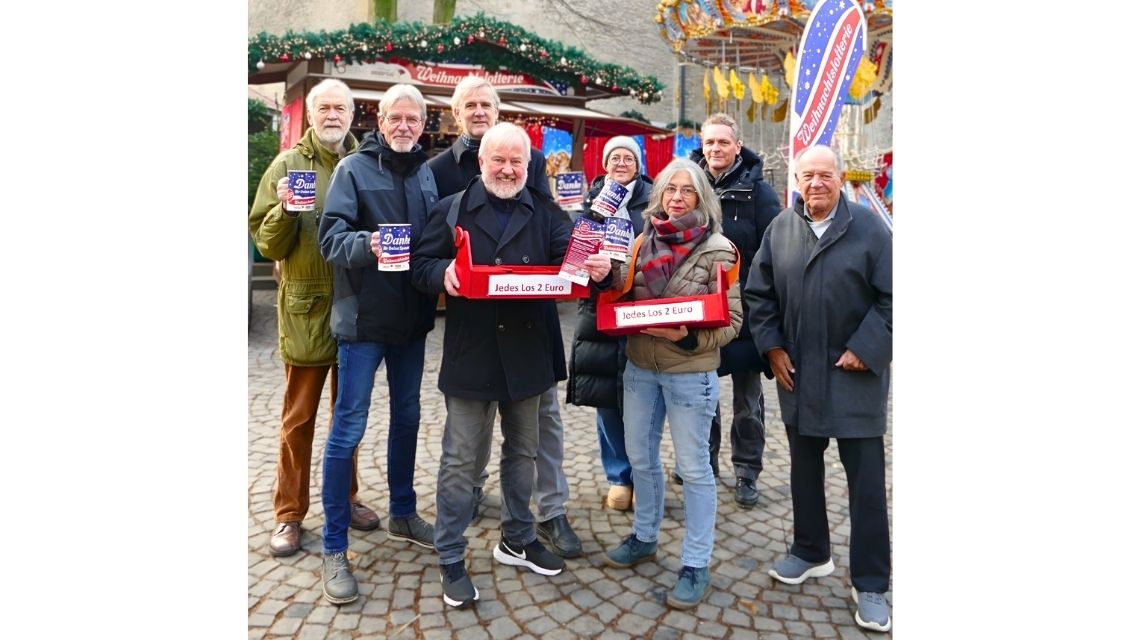 Eine Gruppe Menschen steht vor einem Stand auf dem Weihnachtsmarkt und hält Bauchläden, in denen sich Lotterielose befinden, in ihren Händen.