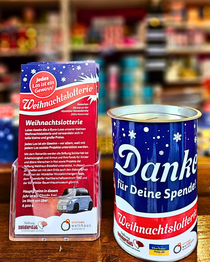 Der Flyer zu der Weihnachtslotterie steht links im Bild, die Spendendose dazu rechts daneben; beides steht auf dem Theresen des Lotteriestandes
