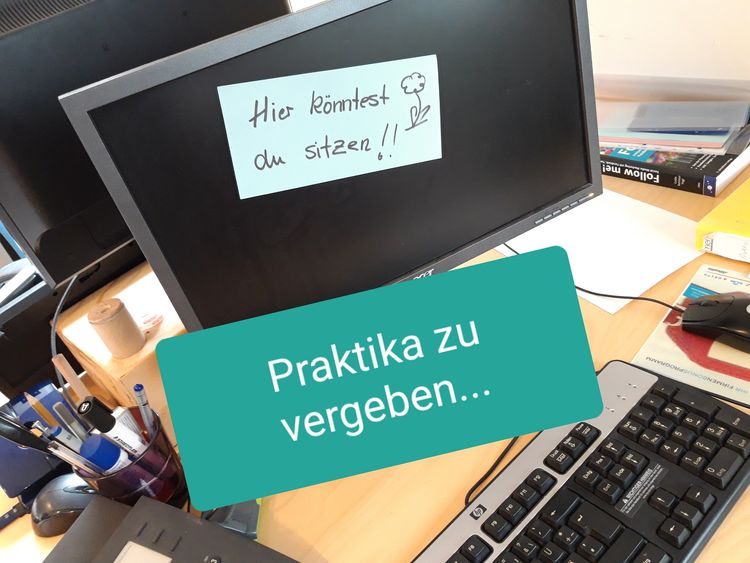 Blick von halboben auf einen Schreibtisch mit Computerbildschirm, auf dem ein Post-it klebt mit dem Text "Hier könntest du sitzen".